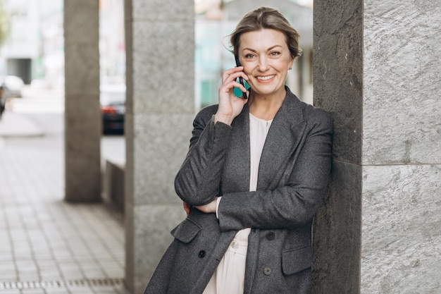 Ritratto di una bella donna d'affari matura sorridente e parlando al telefono sullo sfondo urbano moderno