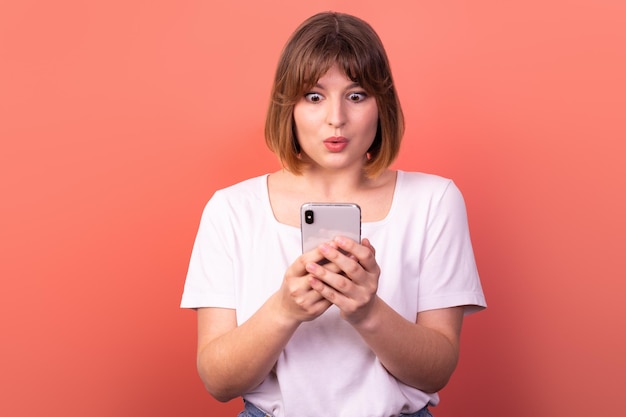 Ritratto di una bella donna dai capelli castani che usa un telefono come reazione WOW su sfondo rosa