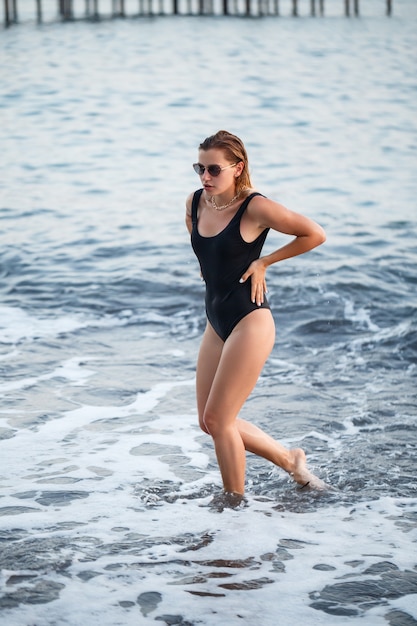 Ritratto di una bella donna in costume da bagno nero con capelli biondi in posa sulla spiaggia in riva al mare. Giovane donna che cammina sulla spiaggia