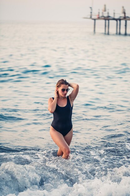 Ritratto di una bella donna in costume da bagno nero con capelli biondi in posa sulla spiaggia in riva al mare Giovane donna che cammina sulla spiaggia