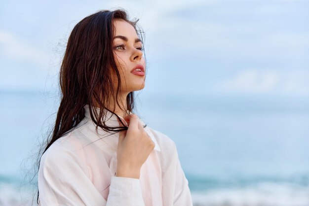 Ritratto di una bella donna pensierosa con la pelle abbronzata in una camicia da spiaggia bianca con i capelli bagnati dopo aver nuotato sulla luce del tramonto dell'oceano sulla spiaggia con le nuvole