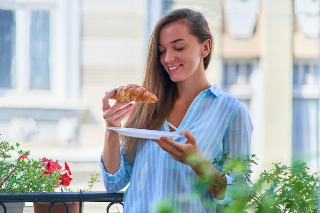 Ritratto di una bella donna romantica sorridente gioiosa carina felice con croissant appena sfornato per la colazione francese al mattino su un balcone