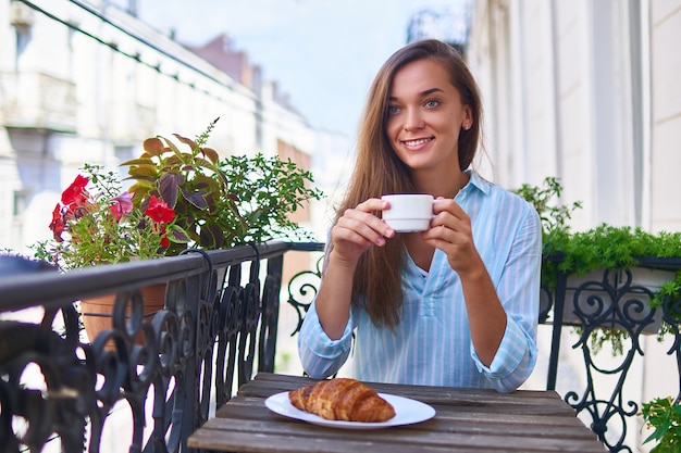 Ritratto di una bella donna romantica sorridente gioiosa carina felice con una tazza di caffè aromatica nelle mani e un piatto di croissant appena sfornato sul tavolo la mattina su un balcone