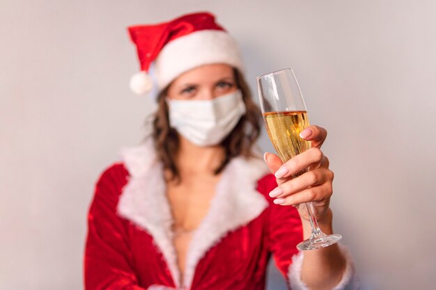 Ritratto di una bella donna sorridente con cappello natalizio e vestito che tiene in mano un bicchiere di champagne mentre si trova su uno sfondo grigio. La donna con la mascherina protettiva dal virus, Natale in quarantena