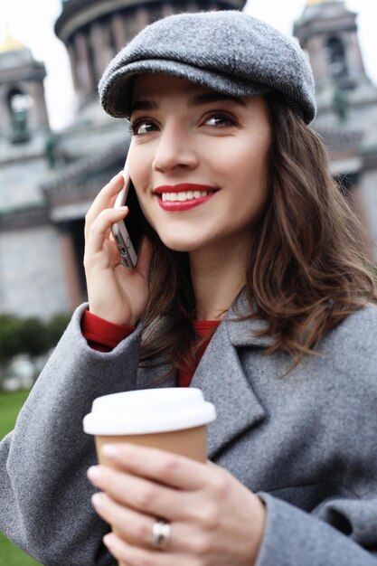 Ritratto di una bella donna sorridente utilizzando il telefono cellulare mentre si tiene la tazza di caffè su una strada cittadina