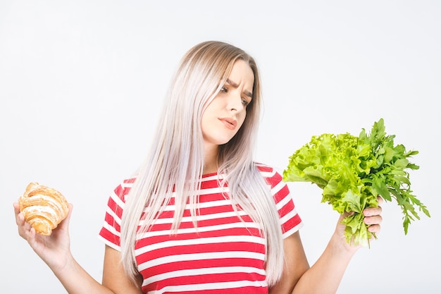 Ritratto di una bella giovane donna bionda confusa scegliendo tra un cibo sano e malsano. Isolato su sfondo bianco.