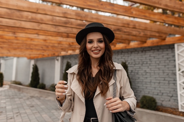 Ritratto di una bella giovane donna bruna riccia con un sorriso carino in un cappello alla moda con un elegante cappotto vintage e una borsa in pelle cammina per strada. Stile femminile elegante, moda e bellezza