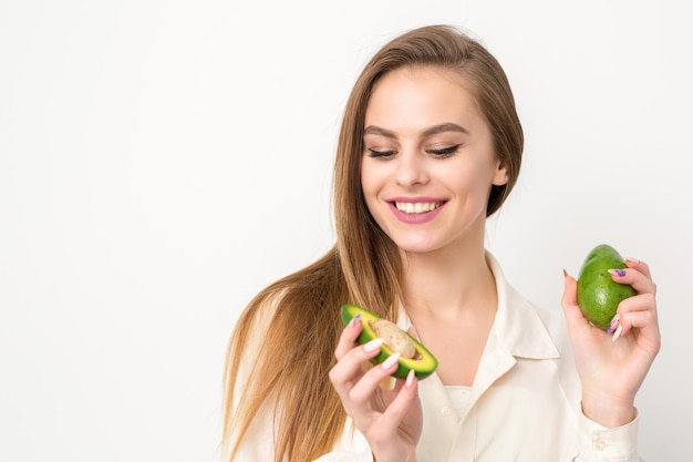 Ritratto di una bella giovane donna caucasica bruna sorridente che indossa la camicia bianca con i capelli lunghi che tengono e mostrano l'avocado in piedi isolato su sfondo bianco