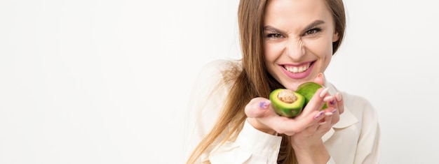 Ritratto di una bella giovane donna caucasica bruna sorridente che indossa la camicia bianca con i capelli lunghi che tengono e mostrano l'avocado in piedi isolato su sfondo bianco