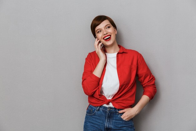 Ritratto di una bella giovane donna che sorride e parla al cellulare isolato su un muro grigio