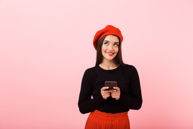 Ritratto di una bella giovane donna felice che indossa in piedi berretto rosso isolato su sfondo rosa, tenendo il telefono cellulare