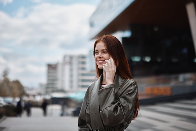 Ritratto di una bella giovane donna in città con un telefono sorriso con denti con capelli rossi volanti in abiti alla moda sullo sfondo della città stile di vita in città