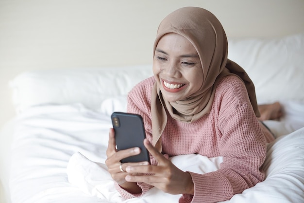 Ritratto di una bella giovane donna musulmana asiatica che indossa l'hijab sul letto che tiene il telefono cellulare