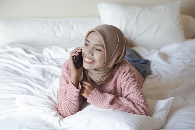 Ritratto di una bella giovane donna musulmana asiatica che indossa l'hijab sul letto che tiene il telefono cellulare