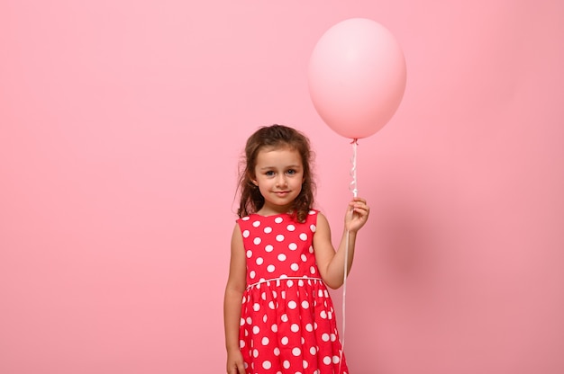 Ritratto di una bella ragazza adorabile di compleanno di 4 anni bella piuttosto splendida, bambino in abito con motivo a pois, con in mano un palloncino rosa, isolato su sfondo rosa con copia spazio per la pubblicità.