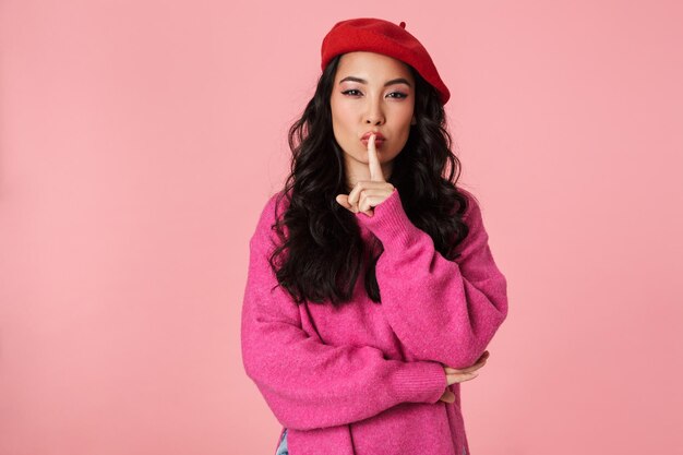 Ritratto di una bella ragazza asiatica sicura di sé che indossa un berretto tenendo il dito indice sulle labbra e chiede di mantenere il silenzio isolato sul rosa
