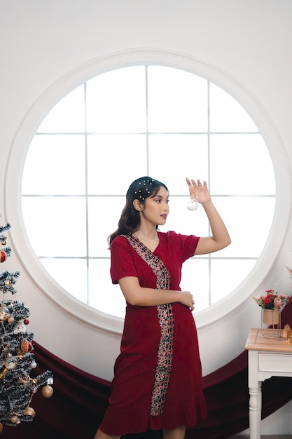 Ritratto di una bella ragazza che indossa un abito rosso che sorride alla macchina fotografica in piedi nel soggiorno decorato di Natale al chiuso