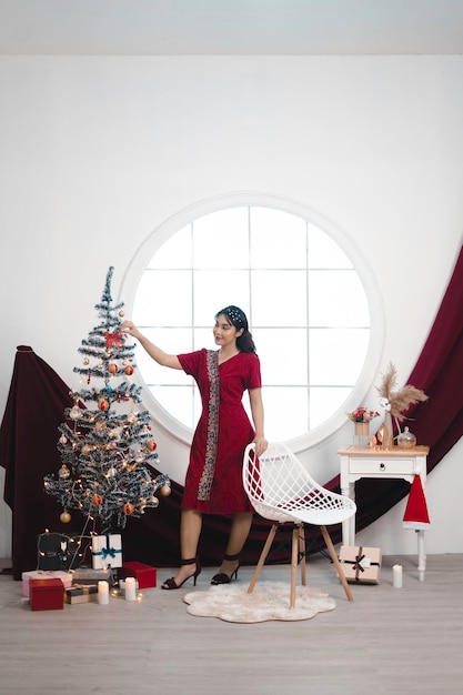 Ritratto di una bella ragazza che indossa un abito rosso che sorride alla macchina fotografica in piedi nel soggiorno decorato di Natale al chiuso