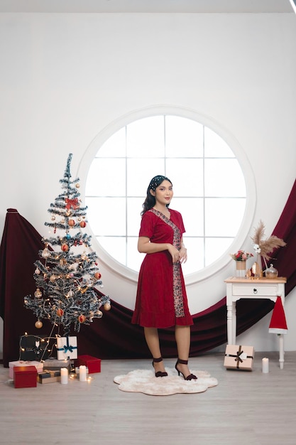Ritratto di una bella ragazza che indossa un abito rosso che sorride alla macchina fotografica in piedi nel soggiorno decorato di Natale al chiuso