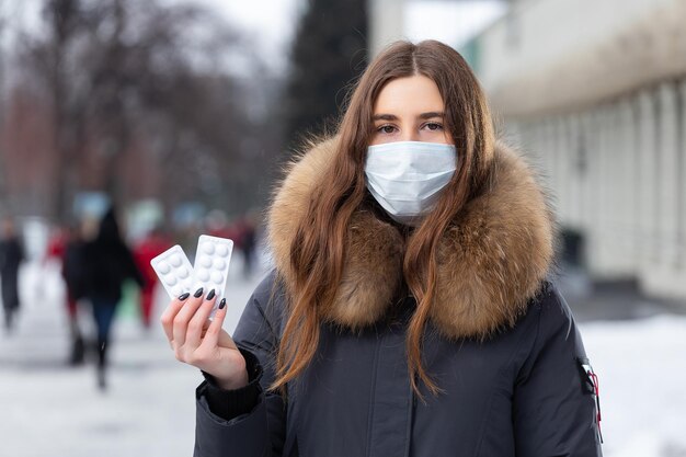 Ritratto di una bella ragazza con una maschera protettiva medica che tiene in mano pillole bianche per raffreddore e influenza Ritratto di strada invernale di una donna sotto la nevicata