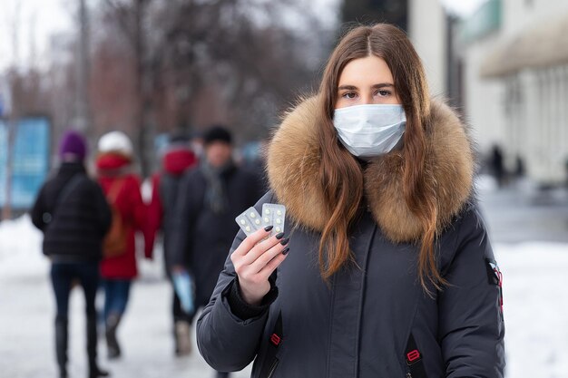 Ritratto di una bella ragazza con una maschera protettiva medica che tiene in mano pillole gialle per raffreddore e influenza Ritratto di strada invernale di una donna sotto la nevicata