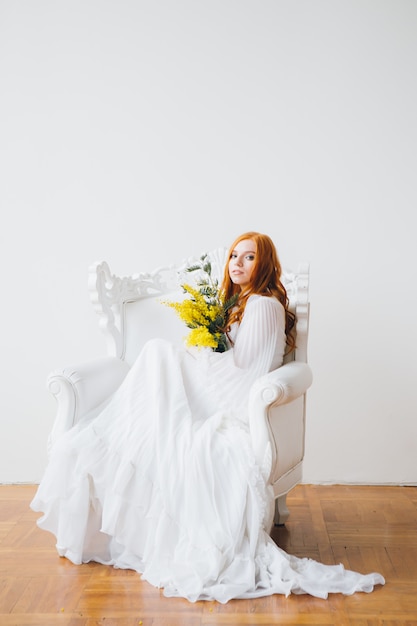 Ritratto di una bella ragazza dai capelli rossi con una mimosa in un lungo abito bianco