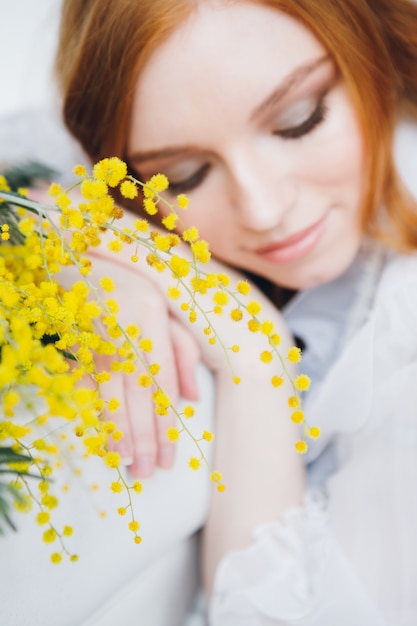 Ritratto di una bella ragazza dai capelli rossi con una mimosa in un lungo abito bianco