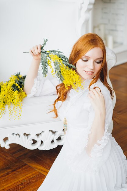 Ritratto di una bella ragazza dai capelli rossi con una mimosa in un lungo abito bianco