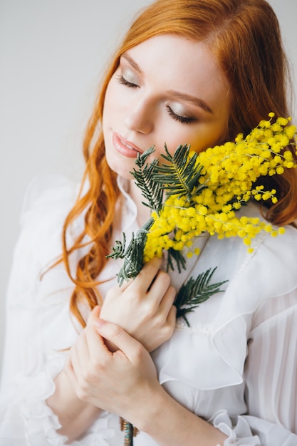 Ritratto di una bella ragazza dai capelli rossi con una mimosa in un lungo abito bianco