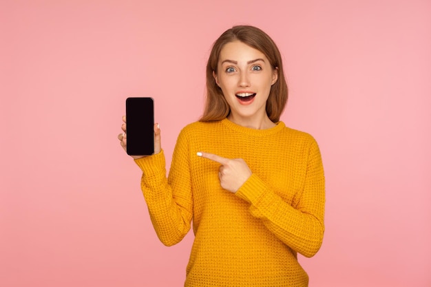 Ritratto di una bella ragazza di zenzero stupita in maglione che punta al cellulare e guarda con sorpresa la fotocamera, scioccata dal dispositivo o dall'applicazione mobile. studio indoor girato sfondo rosa