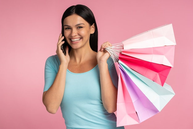 Ritratto di una bella ragazza eccitata che indossa abiti casual chiacchierando allo smartphone mentre tiene in mano le borse della spesa isolate su sfondo rosa