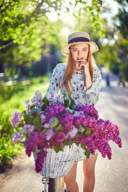 Ritratto di una bella ragazza giovane felice con bicicletta d'epoca e fiori sullo sfondo della città alla luce del sole all'aperto. Bici con cesto pieno di fiori. Concetto di tempo libero attivo.