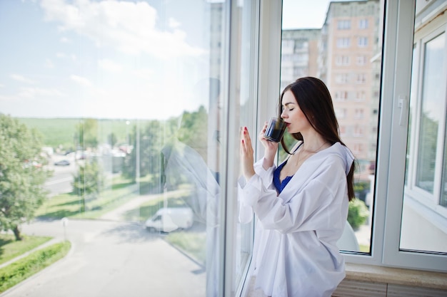 Ritratto di una bella ragazza in biancheria intima e camicia maschile in piedi con un bicchiere d'acqua in mano sul balcone