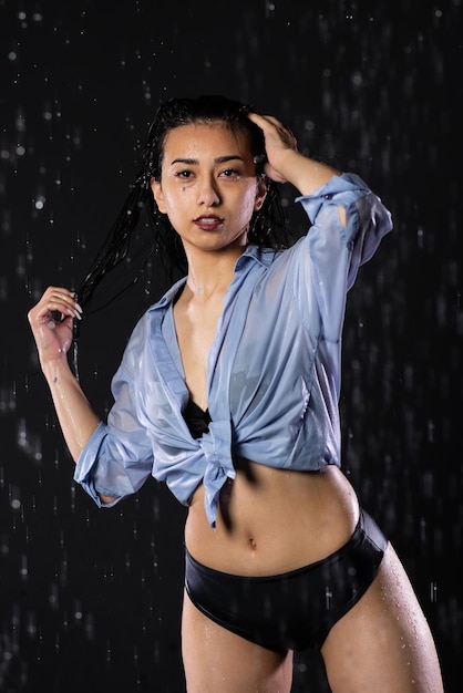Ritratto di una bella ragazza in un costume da bagno nero e camicia In piedi sotto la pioggia godendo le gocce d'acqua Spruzzi d'acqua su sfondo nero Studio Aqua