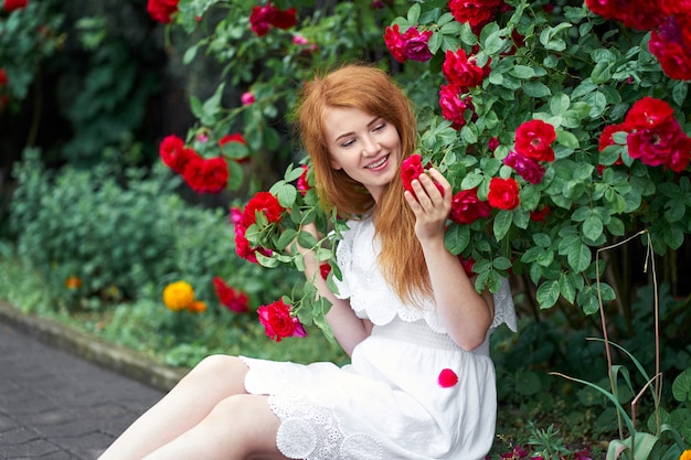 Ritratto di una bella ragazza rossa vestita con un abito a luce bianca su uno sfondo di rose in fiore.