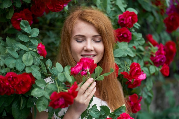 Ritratto di una bella ragazza rossa vestita con un abito a luce bianca su uno sfondo di rose in fiore.