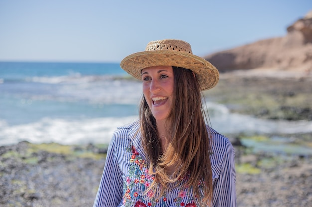 Ritratto di una bella ragazza sorridente in spiaggia, che indossa una camicia a fiori e un cappello estivo.