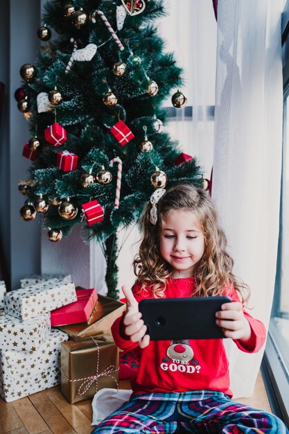 Ritratto di una bella ragazzina a casa vicino all'albero di Natale e usando il telefono cellulare