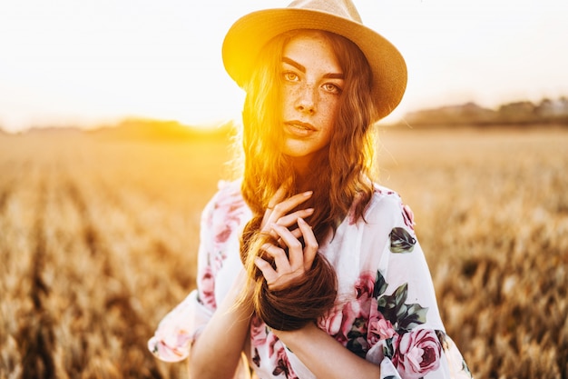 Ritratto di una bellissima giovane donna con i capelli ricci e le lentiggini faccia. Donna in vestito e cappello che posano nel campo di frumento al tramonto e allo sguardo