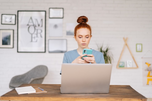 Ritratto di una bellissima giovane donna dai capelli rossi che usa il telefono cellulare mentre è seduta alla scrivania con un laptop