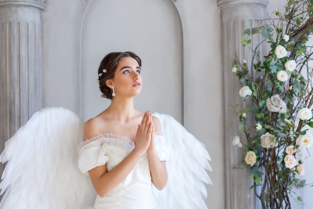 Ritratto di una bellissima giovane donna in abito bianco e ali d'angelo, in piedi con uno sguardo supplichevole sullo sfondo di un muro con colonne e fiori