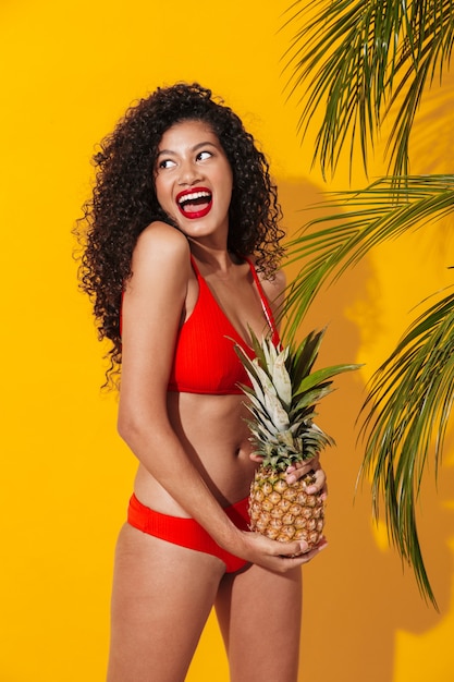 Ritratto di una bellissima giovane donna in forma felice che indossa un bikini in piedi isolato su una parete esotica gialla, in posa con ananas