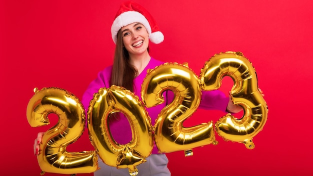 Ritratto di una bruna ridente che detiene i numeri del nuovo anno 2023 Una giovane donna indossa un cappello di Babbo Natale