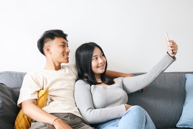 Ritratto di una coppia asiatica felice seduta sul divano e che si fa un selfie insieme con lo smartphone a casa