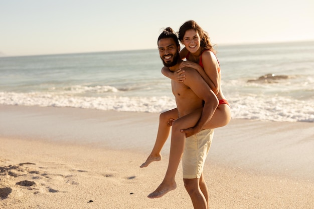 Ritratto di una coppia caucasica abbronzata in vacanza, che si diverte a fare il giro sulle spalle su una spiaggia assolata, guardando la macchina fotografica e sorridendo, con cielo blu e mare sullo sfondo