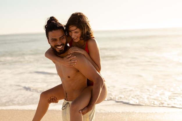 Ritratto di una coppia caucasica abbronzata in vacanza, che si diverte a fare il giro sulle spalle su una spiaggia assolata, guardando la macchina fotografica e sorridendo, con cielo blu e mare sullo sfondo