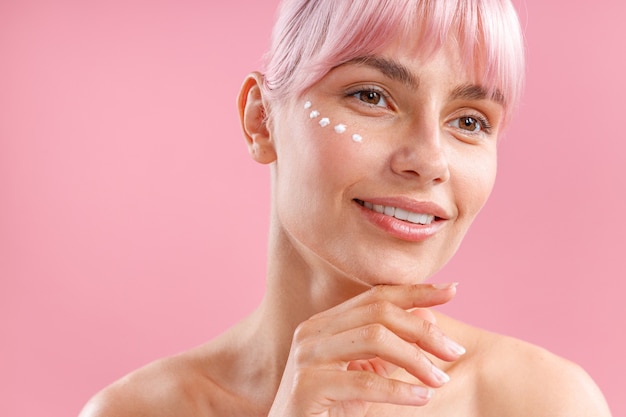 Ritratto di una donna adorabile con i capelli rosa e la crema per il viso applicati sulla pelle come puntini che sorridono via