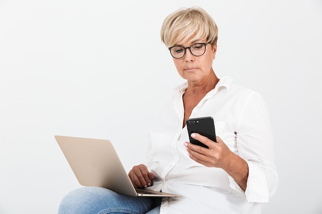 Ritratto di una donna adulta attraente che indossa occhiali che tiene lo smartphone mentre è seduto con un computer portatile isolato su un muro bianco