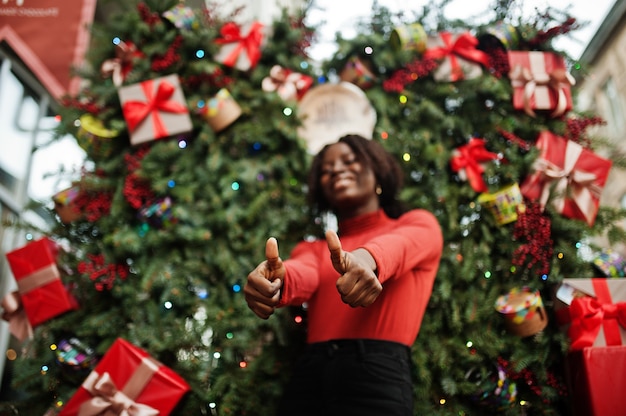 Ritratto di una donna africana dai capelli ricci che indossa dolcevita rosso alla moda in posa contro le decorazioni natalizie, tema della vigilia di capodanno. Mostra pollice in alto.