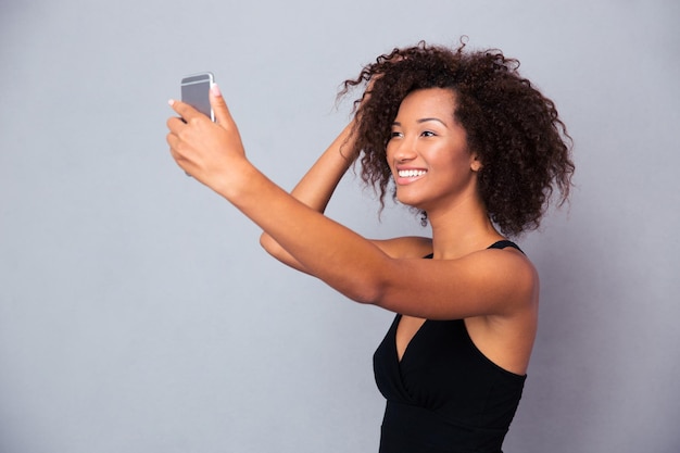 Ritratto di una donna afroamericana sorridente che fa la foto del selfie sullo smartphone sopra la parete grigia